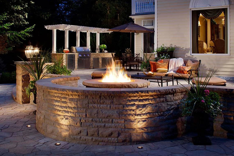 Outdoor kitchen with pergola and firepit with surrounding stone seating, outdoor lighting, ambiance lighting, walkway lighting